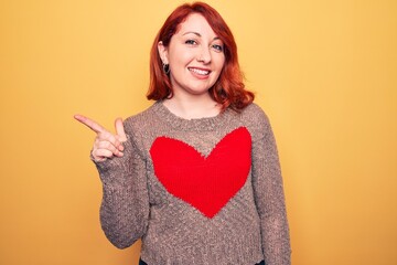 Young beautiful redhead woman wearing casual sweater with heart over yellow background smiling cheerful pointing with hand and finger up to the side