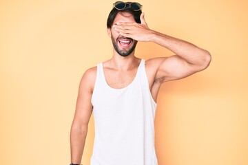 Handsome hispanic man wearing summer look and sunglasses smiling and laughing with hand on face covering eyes for surprise. blind concept.