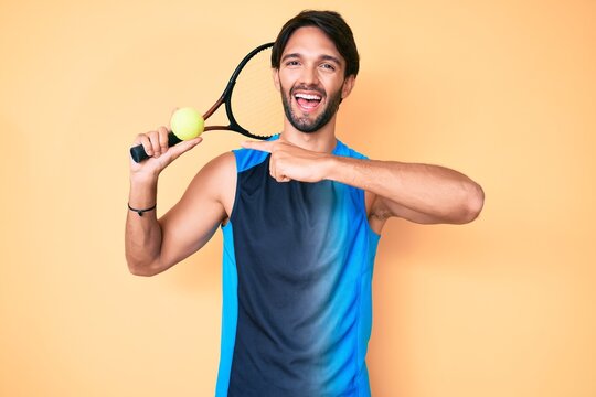 Handsome hispanic man playing tennis holding racket and ball smiling happy pointing with hand and finger