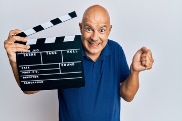 Middle age bald man holding video film clapboard screaming proud, celebrating victory and success...