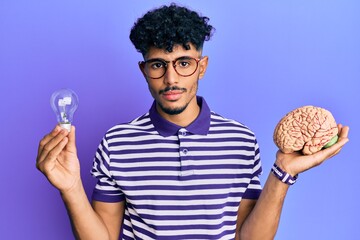 Young arab handsome man holding brain and lightbulb for inspiration and idea relaxed with serious...