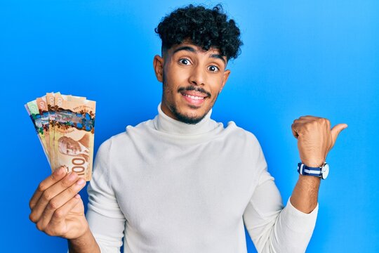 Young arab handsome man holding canadian dollars pointing thumb up to the side smiling happy with open mouth