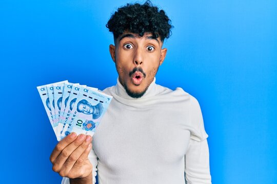 Young arab handsome man holding 10 yuan chinese banknotes scared and amazed with open mouth for surprise, disbelief face