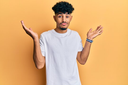 Young arab handsome man wearing casual white tshirt clueless and confused with open arms, no idea and doubtful face.