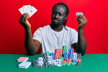 Handsome young black man sitting on the table with poker chips and cards clueless and confused expression. doubt concept.