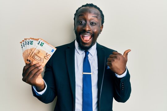 Handsome young black man wearing business suit holding 50 euros banknotes pointing thumb up to the side smiling happy with open mouth