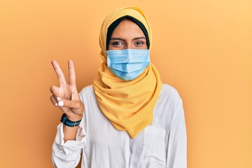 Young brunette arab woman wearing traditional islamic hijab and medical mask smiling with happy face winking at the camera doing victory sign. number two.