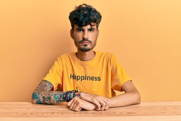Young hispanic man wearing tshirt with happiness word message sitting on the table skeptic and nervous, frowning upset because of problem. negative person.