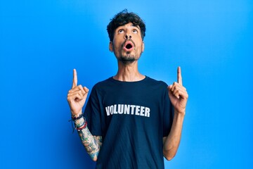 Young hispanic man wearing volunteer t shirt amazed and surprised looking up and pointing with fingers and raised arms.