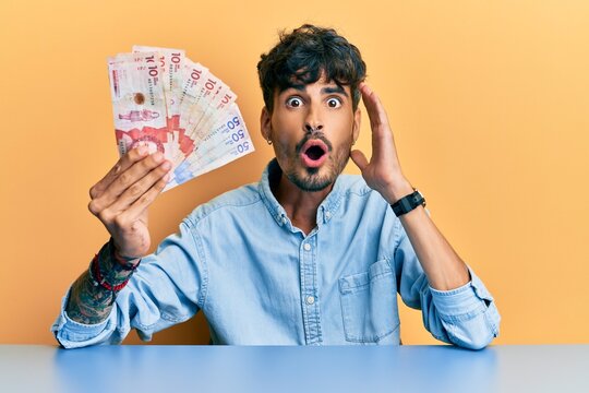 Young hispanic man holding colombian pesos sitting on the table scared and amazed with open mouth for surprise, disbelief face