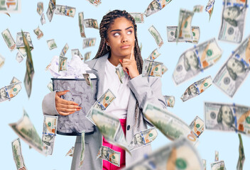 Young african american woman with braids holding paper bin full of crumpled papers serious face thinking about question with hand on chin, thoughtful about confusing idea