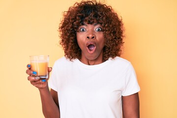 Young african american woman drinking glass of orange juice scared and amazed with open mouth for surprise, disbelief face