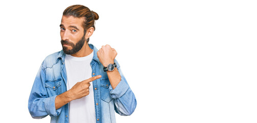 Attractive man with long hair and beard wearing casual denim jacket in hurry pointing to watch...