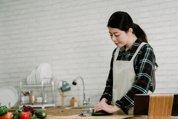 young elegant asian korean woman in modern kitchen following recipe online website on digital tablet. smiling wife in apron chopping cucumber with sharp knife on wooden cutting board in cooking place