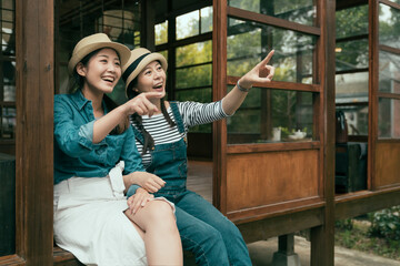 two cheerful young asian chinese girl travelers pointing finger away while relax sitting on wooden floor. friends in straw hats smiling enjoy garden view from japanese traditional design house.