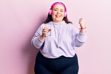 Naklejka premium Young plus size woman drinking glass of water using headphones screaming proud, celebrating victory and success very excited with raised arms