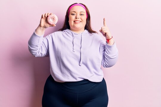 Young plus size woman holding donut smiling with an idea or question pointing finger with happy face, number one