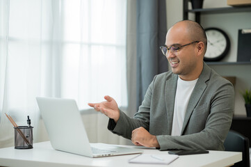 A middle-aged man around the age of 35. Working at home Work through the laptop by meeting video conference. He was wearing a grey suit and glasses. Smiling asian businessman work from home..