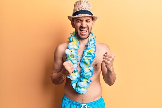 Young handsome bald man shirtless wearing swimwear and hawaiian lei celebrating surprised and amazed for success with arms raised and eyes closed