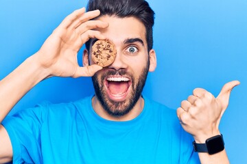 Young handsome man with beard holding chocolate cookie pointing thumb up to the side smiling happy with open mouth