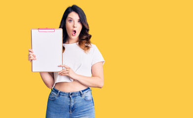 Fototapeta na wymiar Beautiful young brunette woman holding clipboard with blank space scared and amazed with open mouth for surprise, disbelief face