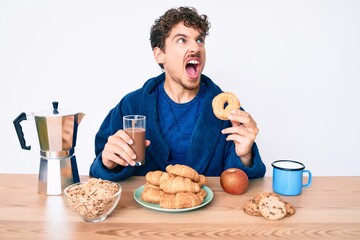 Young caucasian man with curly hair eating breakfast holding chocolate beverage and donut angry and mad screaming frustrated and furious, shouting with anger. rage and aggressive concept.