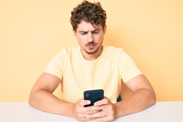 Young caucasian man with curly hair using smartphone sitting on the table thinking attitude and sober expression looking self confident