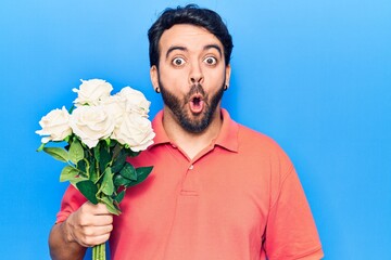 Young hispanic man holding flowers scared and amazed with open mouth for surprise, disbelief face