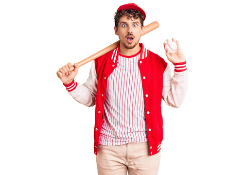Young handsome man with curly hair playing baseball holding bat and ball scared and amazed with open mouth for surprise, disbelief face