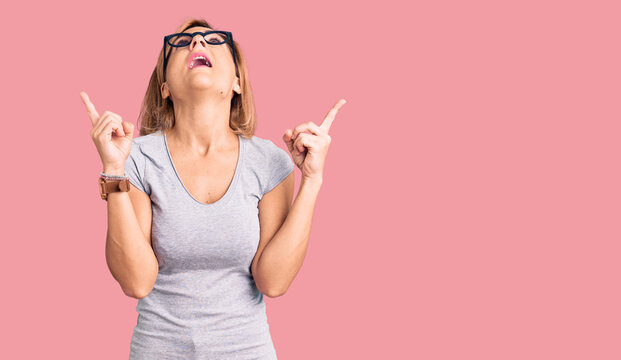 Young blonde woman wearing casual clothes amazed and surprised looking up and pointing with fingers and raised arms.