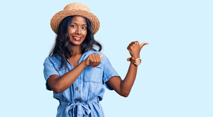 Young indian girl wearing summer hat pointing to the back behind with hand and thumbs up, smiling confident