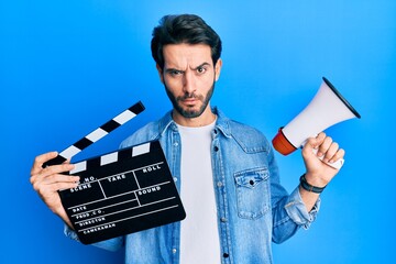 Young hispanic man holding video film clapboard and megaphone skeptic and nervous, frowning upset...