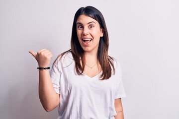 Young beautiful brunette woman wearing casual t-shirt standing over isolated white background pointing thumb up to the side smiling happy with open mouth