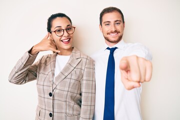 Beautiful couple wearing business clothes smiling doing talking on the telephone gesture and pointing to you. call me.