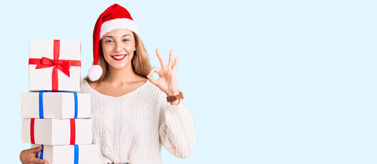 Fototapeta premium Beautiful young woman wearing christmas hat and holding a gift doing ok sign with fingers, smiling friendly gesturing excellent symbol