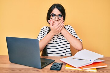 Brunette teenager girl working at the office with laptop shocked covering mouth with hands for mistake. secret concept.
