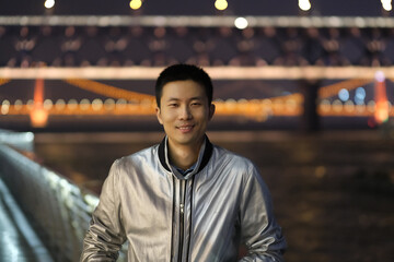 medium shot of one young Asian man looking at camera at night. Smiling happily. Defocused Wuhan...