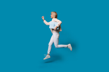 Running blonde woman is holding a present box on a blue studio wall and smile in casual clothes