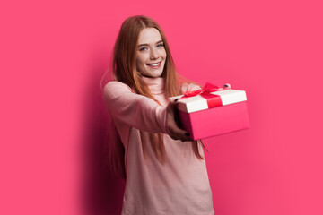 Fototapeta premium Ginger woman with freckles and red hair is smiling and giving at camera a red box with gift posing on a red wall