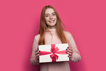 Caucasian woman with red hair and freckles is showing at camera present box well packed posing and smile on a red wall