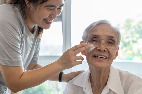 Female caregiver assisting to apply sunscreen lotion on the face of senior woman,granddaughter using skin care cream for elderly sensitive skin,happy smiling grandmother with sun block on facial skin