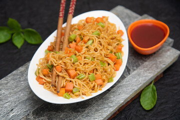 Schezwan Noodles or vegetable Hakka Noodles or chow mein, served in a bowl or plate with tomato sauce, ketchup, wooden chopsticks on dark black background Kerala, India. popular Indo-Chinese recipes.