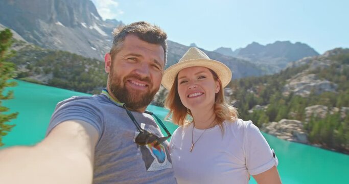 Romantic couple kissing in camera with scenic lake and mountain landscape on background. Man and woman loving each other taking selfie picture or video on sunny summer day. 4K happy people travel USA