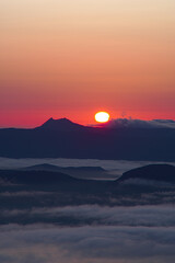 Fototapeta na wymiar 遠くの山の上に昇る朝の太陽。薄明りの地上の風景。オレンジ色の夜明けの空。