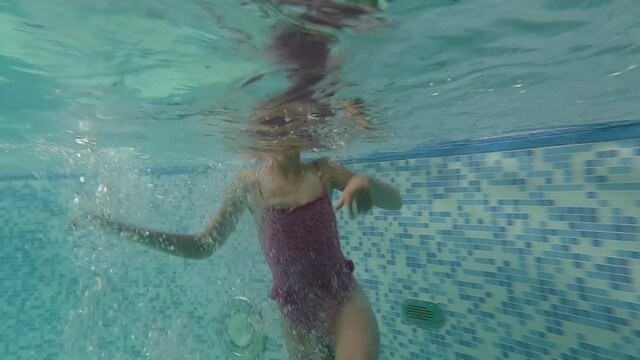 Underwater shooting of a funny little girl swimming in a large pool in glasses and a pink bikini. The child vigorously moves his arms and legs and blows bubbles