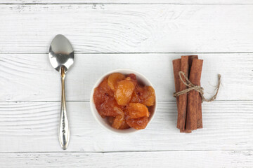 bowl of apple jam, spoon and cinnamon sticks on white wooden background flat lay