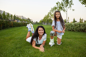 Fototapeta premium mother and daughter in roller skates are sitting on grass and having fun in park.