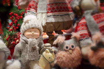 Cute baby doll in a winter costume on a horse, knitted hat and scarf. Rag fluffy owl and other Christmas toys gifts. Blurred background, christmas decoration in toy store