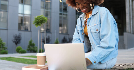 Young African American curly beautiful woman sitting on bench, working on laptop computer and...