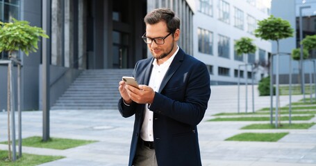 Caucasian happy young stylish businessman tapping or scrolling on smartphone and standing at street. Handsome male texting message on phone and smiling. Outdoors.